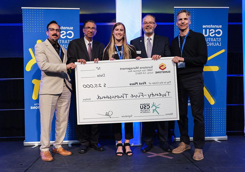 A female, college-age startup founder poses onstage with two sponsors, two hosts, and a giant check for $25,000