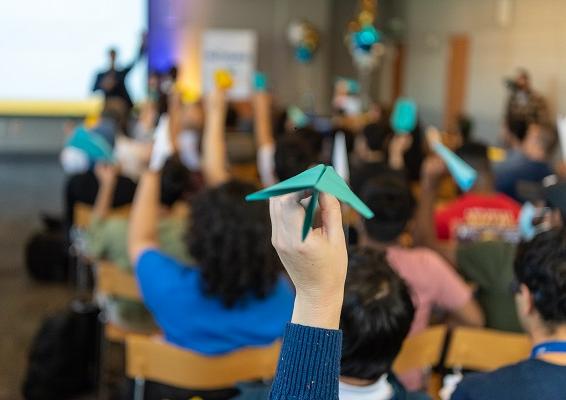 A roomful of people, seen from the back, hold up blue, white and gold paper planes, poised to throw toward an out-of-focus speaker at the front of the room 